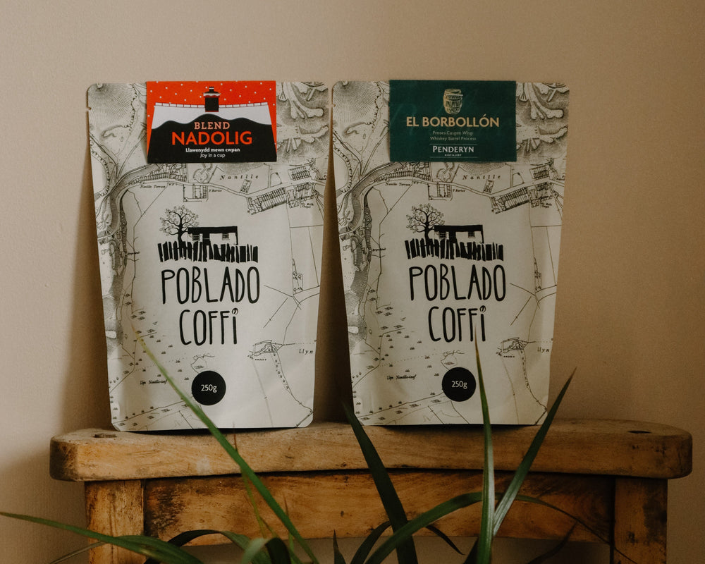 Two coffee bags labeled 'Poblado Coffee' on a wooden stool with a plant in the foreground.